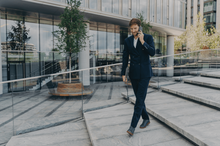 Un homme en costume sombre descend des marches devant un bâtiment moderne en verre, tout en parlant au téléphone. Cette image illustre parfaitement le concept de téléphonie professionnelle, montrant un professionnel en déplacement, utilisant son téléphone pour des communications d'affaires.