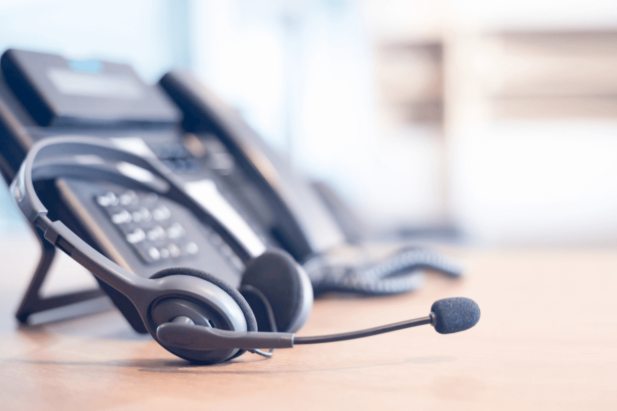 Cette image montre un casque téléphonique avec microphone posé sur une table, à côté d'un téléphone de bureau. Cette scène est typique de la téléphonie professionnelle, où les employés utilisent des casques pour gérer les appels de manière efficace et confortable.